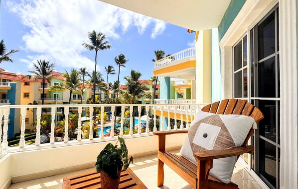 A balcony with a wooden chair and a potted plant, overlooking a pool and resort buildings.