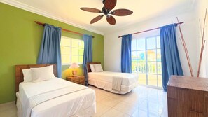 Une chambre d’hôtel avec deux lits, un ventilateur de plafond et de grandes fenêtres avec des rideaux bleus.