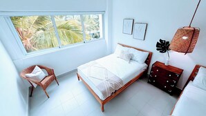 Een slaapkamer met een bed, een stoel, een nachtkastje, een lamp en een raam met uitzicht op palmbomen.