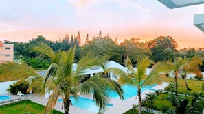 A pool area with palm trees, a building, and a sunset in the background.