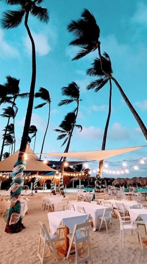A beachside restaurant with outdoor seating, palm trees, and string lights.