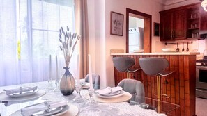 A dining area with a table set for four, a vase with dried flowers, and a kitchen in the background.