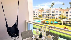 Un balcón con una hamaca colgante, una mesa con un calentador de agua, tazas y una planta pequeña, con vistas a una piscina y un edificio de resort.