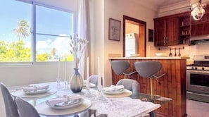 Uma área de jantar com mesa posta para quatro pessoas, um vaso com flores e vista para palmeiras lá fora.