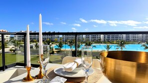 A table set with a white plate, a glass, a gold bowl, and two candles on a balcony overlooking a pool and hotel buildings.