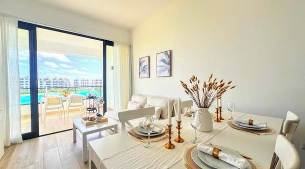 A dining area with a table set for four, a balcony with a view of buildings and a pool, and two framed pictures on the wall.