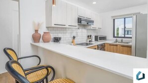 A modern kitchen with white cabinets, a white countertop, and a stainless steel refrigerator. There are two wicker chairs and a small table.