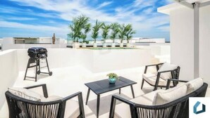 A rooftop terrace with outdoor furniture, a grill, and palm trees.