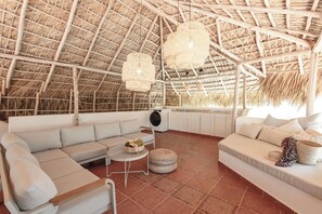 Une pièce spacieuse avec un toit de chaume, un ensemble de canapés blancs, un pouf rond en osier et une petite table sur laquelle se trouve un bol.