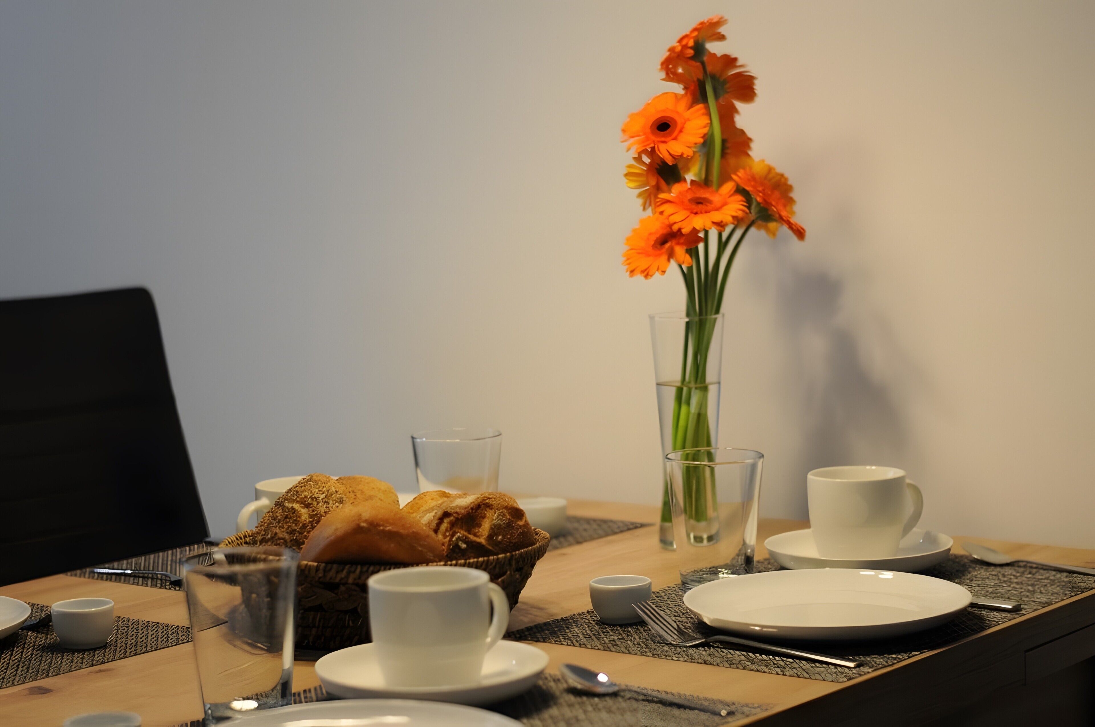 Een eettafel gedekt met borden, kopjes, een mand met brood, een vaas met bloemen en een glas water.