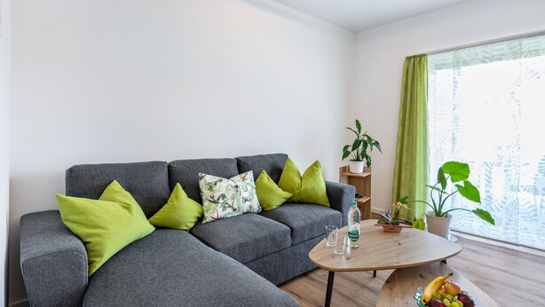 A modern living room with a grey sectional sofa, green cushions, a wooden coffee table, and a potted plant.