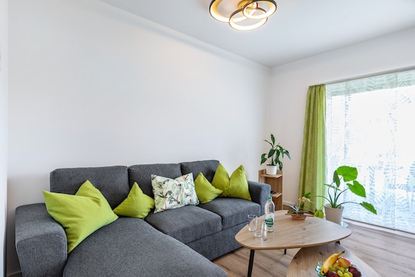 Ein modernes Wohnzimmer mit einem grauen Ecksofa, grünen Kissen, einem Holztisch und einer Zimmerpflanze.