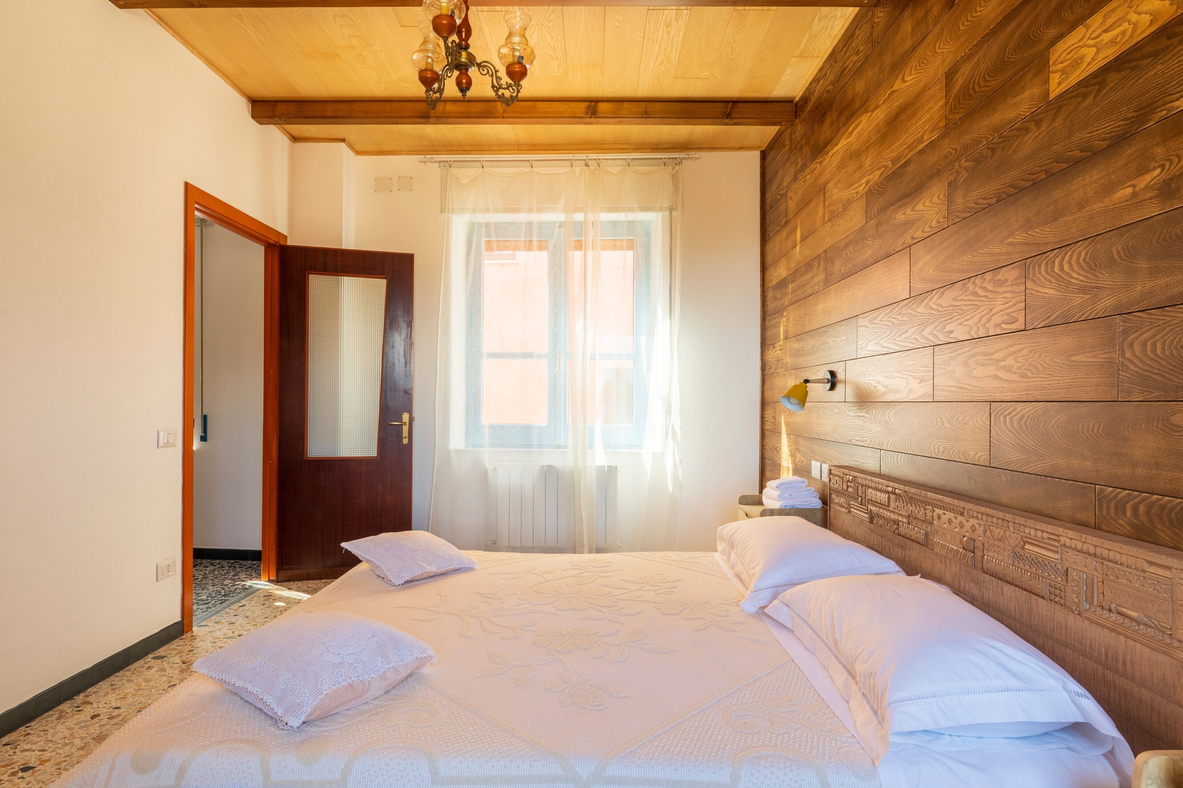 Ein Schlafzimmer mit einem hölzernen Kopfteil, einem Bett mit weißer Bettwäsche, einem Fenster mit durchscheinenden Vorhängen und einer Pendelleuchte.