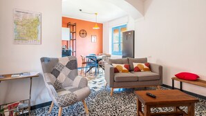 Un salon comprenant un canapé, un fauteuil et une table basse, un coin cuisine avec un réfrigérateur, et un espace repas avec des chaises et une table.