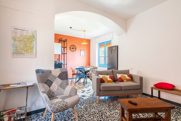 A living room with a sofa, armchair, and coffee table, a kitchen area with a fridge, and a dining space with chairs and a table. - Holiday Home 'Sa Omu 'Eccia' with Sea View and Private Terrace (Villagrande Strisaili)