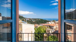 Une vue à travers une fenêtre donnant sur un balcon, avec en contrebas une ville côtière, des bâtiments et, au loin, une montagne.