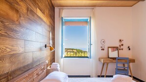 Une chambre avec une tête de lit en bois, un lit avec du linge de maison blanc, un bureau avec une chaise, une fenêtre donnant sur un beau paysage et un petit balcon.