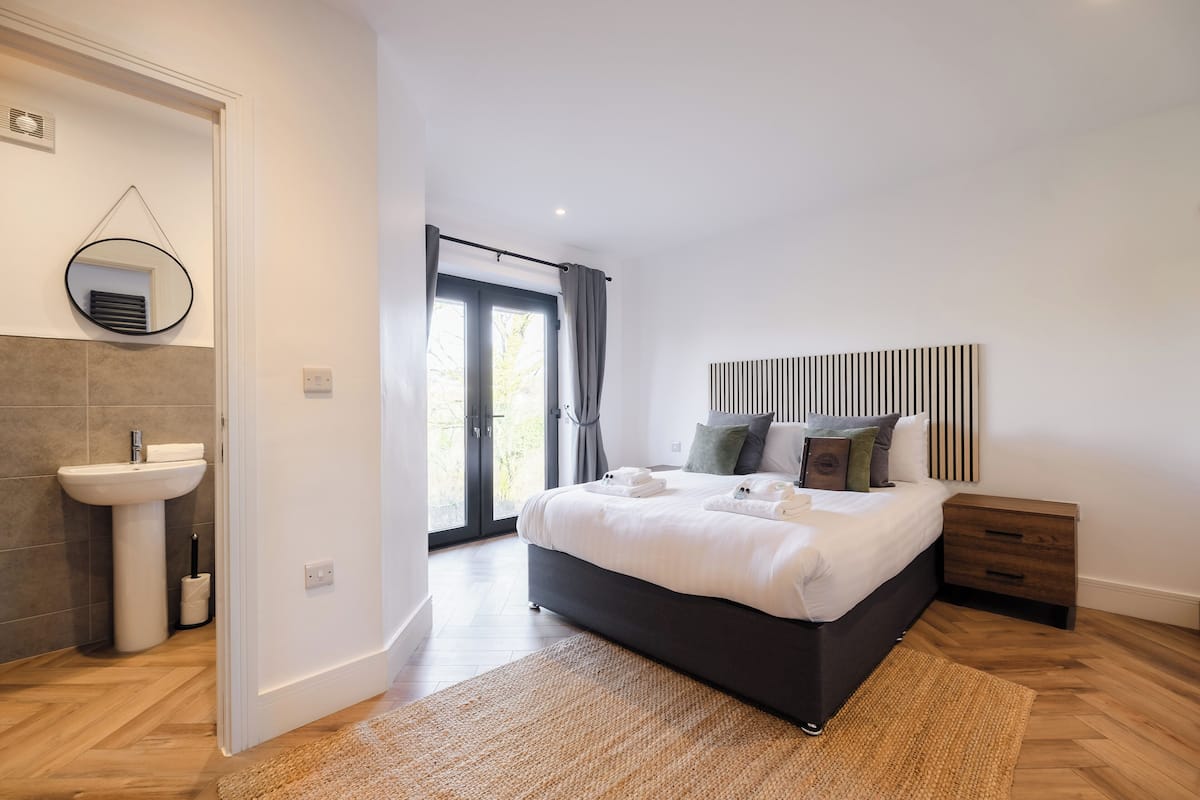 A modern bedroom with a large bed, bedside tables, a round mirror, and a view through a glass door.