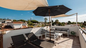 Una terraza en la azotea con un sofá, dos sillones, una mesa y un amplio paraguas.