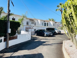 Una calle residencial con coches estacionados, un edificio blanco con techo de tejas y una planta de cactus.