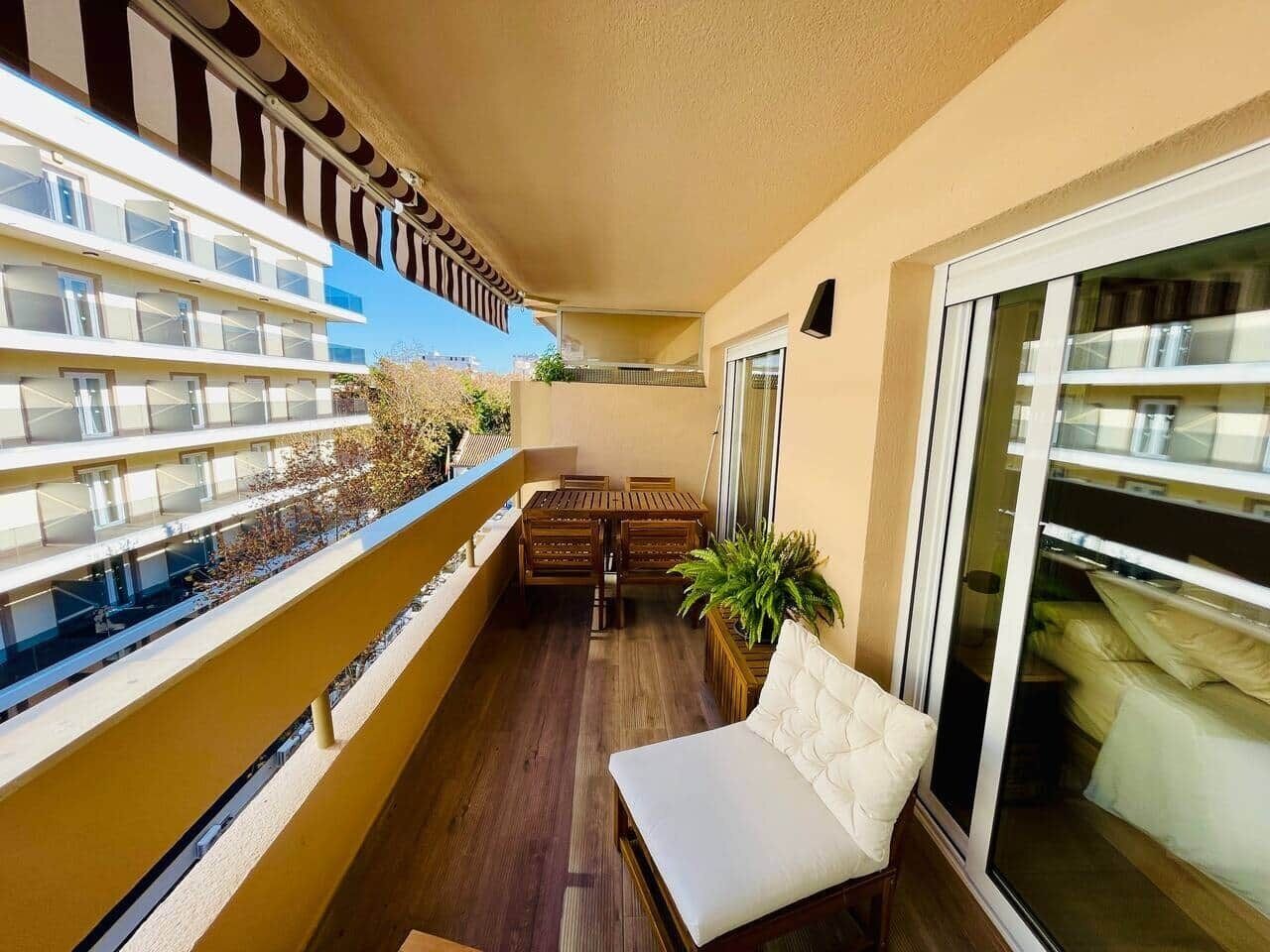 A balcony with a wooden table and chairs, a cushioned bench, and potted plants.