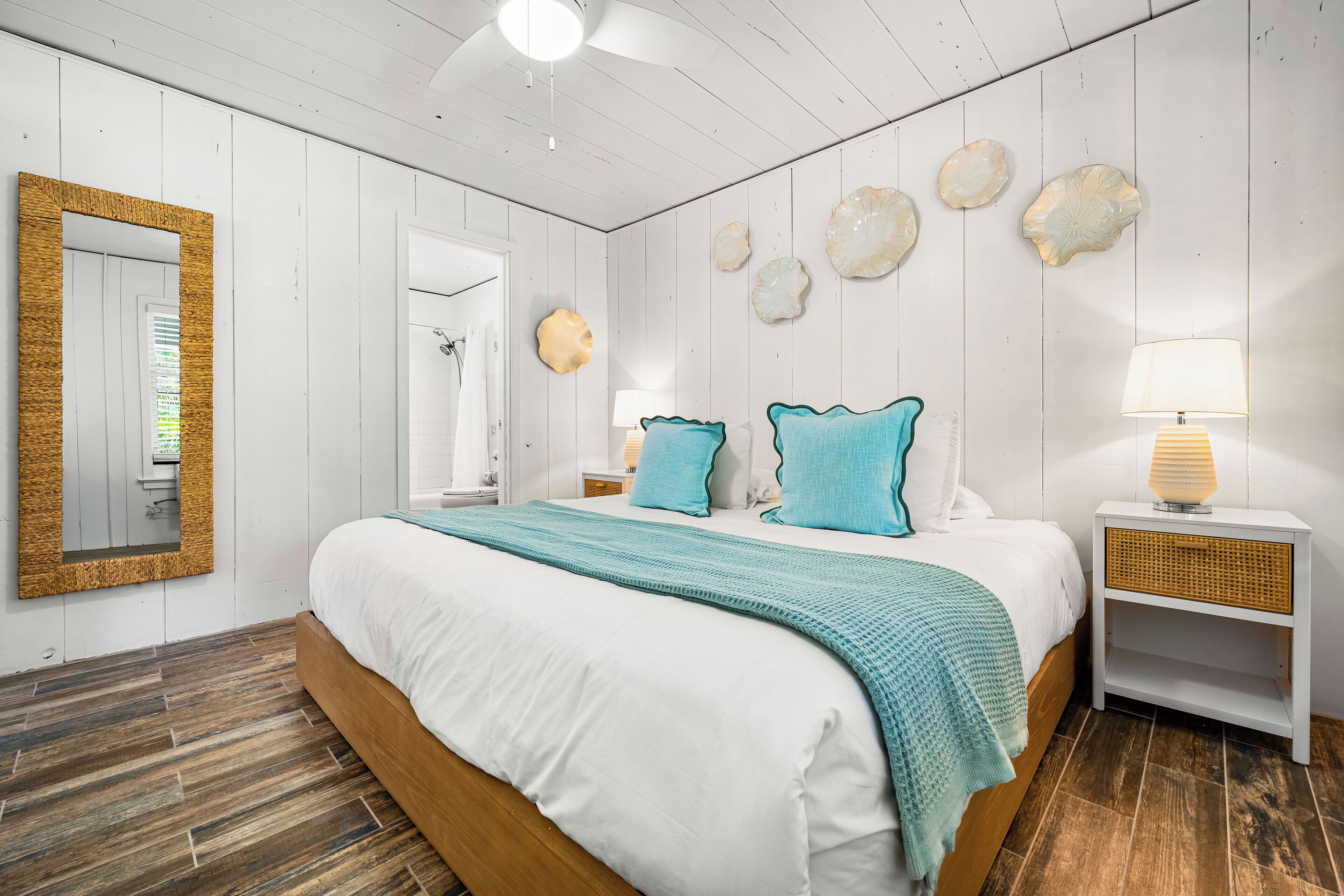 A bedroom with a wooden bed, white bedding, teal throw pillows, a white nightstand with a lamp, and decorative shells on the wall.