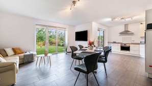 A modern kitchen with a flat-screen TV, a dining area with a table and chairs, and a sofa with a view of the outdoors. - Holiday Home 'Großes Im Grünen -' with Private Terrace, Private Garden and Wi-Fi (Dummerstorf)