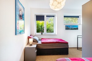 Ein Schlafzimmer mit Bett, Nachttisch, Lampe, zwei Fenstern und einem Wandbild.