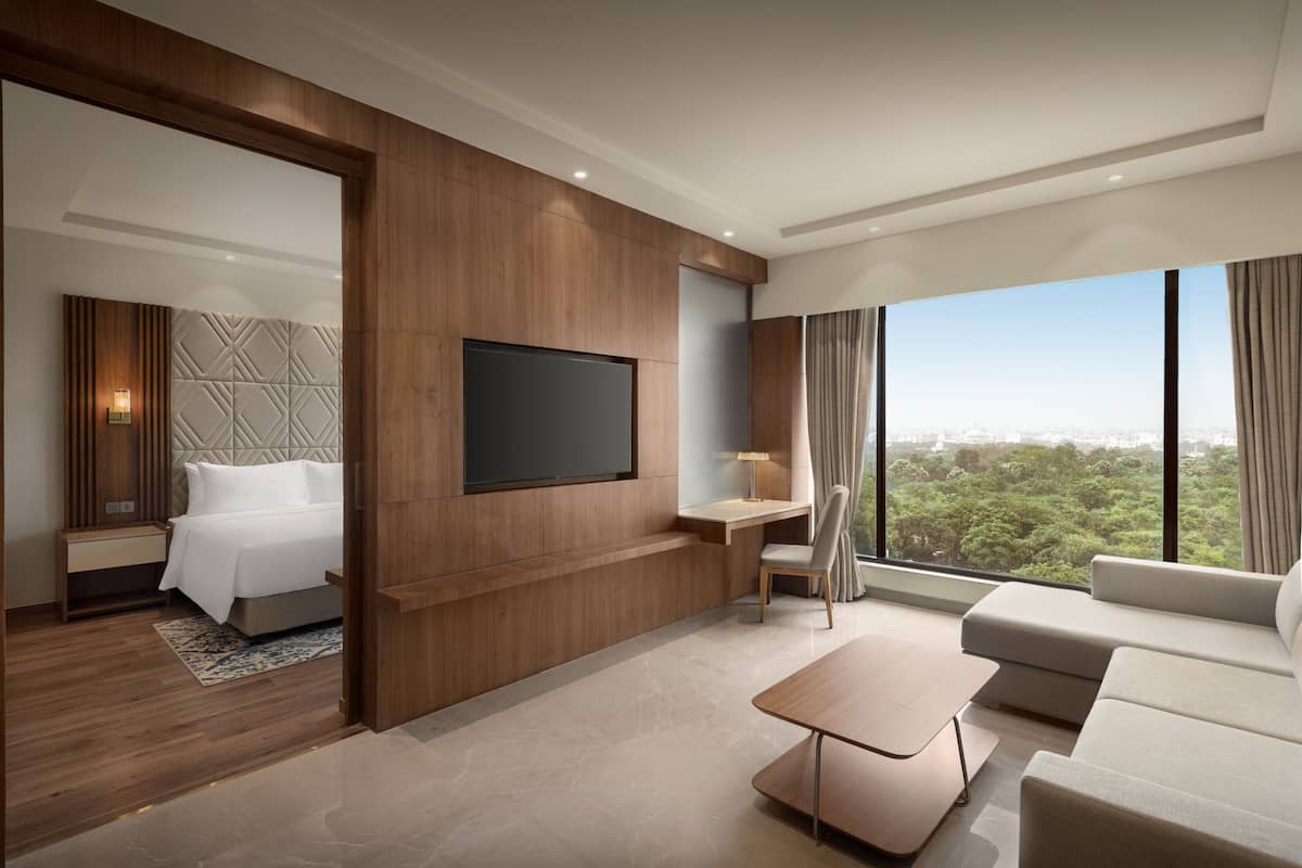 A modern hotel room with a large bed, a flat-screen TV mounted on the wall, a seating area with a sofa and a coffee table, and a large window offering a city view.