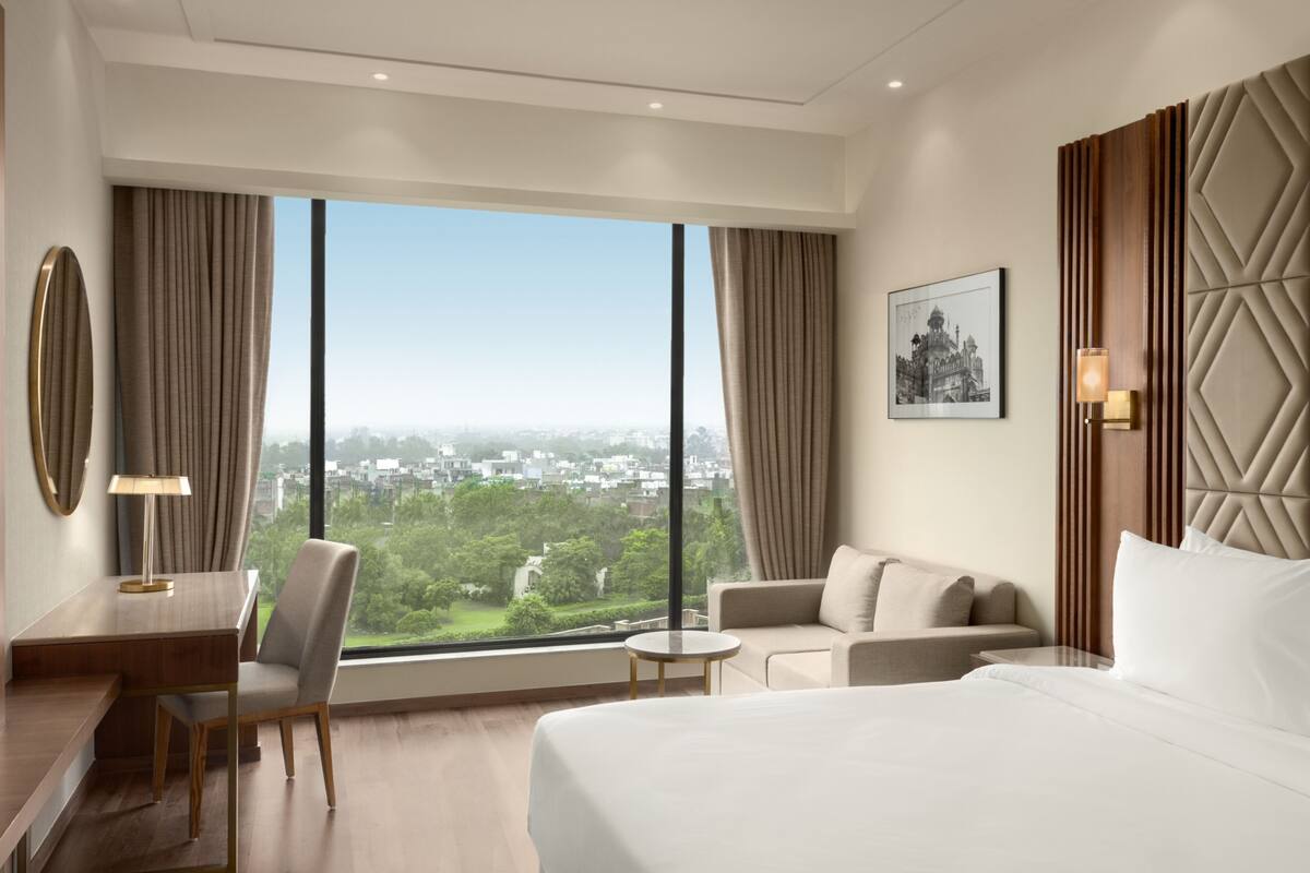 A hotel room with a large window, a bed, a desk, a chair, and a sofa.