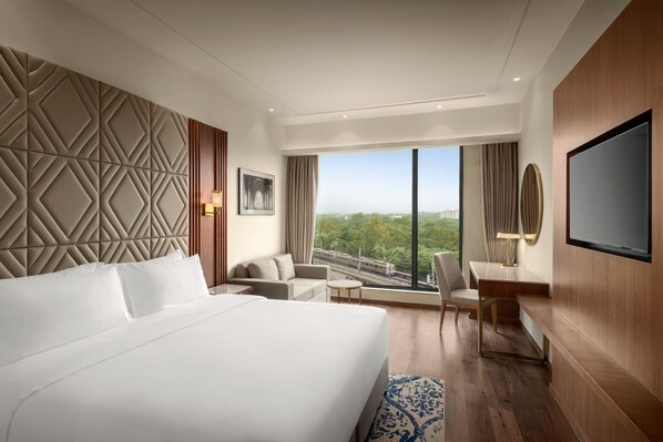A modern hotel room with a large bed, a desk, a chair, and a TV. There is a large window with a view of the city. - Radisson Hotel Delhi MG Road (New Delhi)