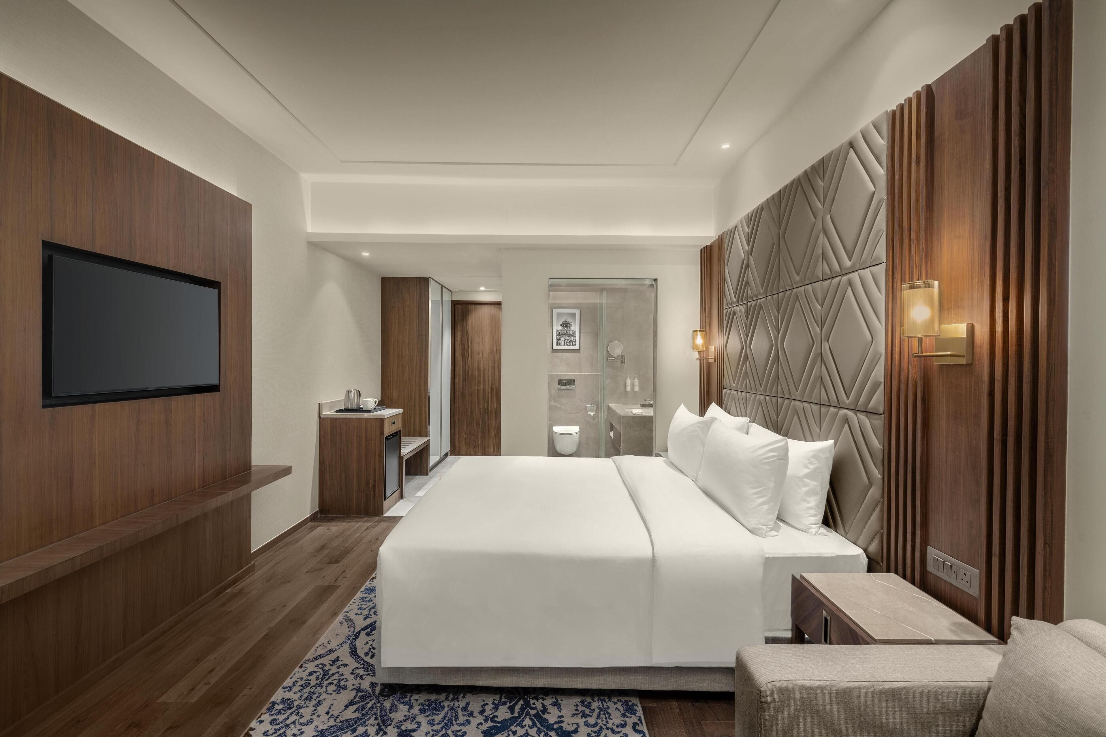 A modern hotel room with a large bed, a TV mounted on the wall, a bedside table, and a wooden headboard with built-in lighting.