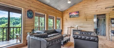 A log cabin with a large living area featuring leather sofas, a fireplace, and large windows offering a view of the forest.