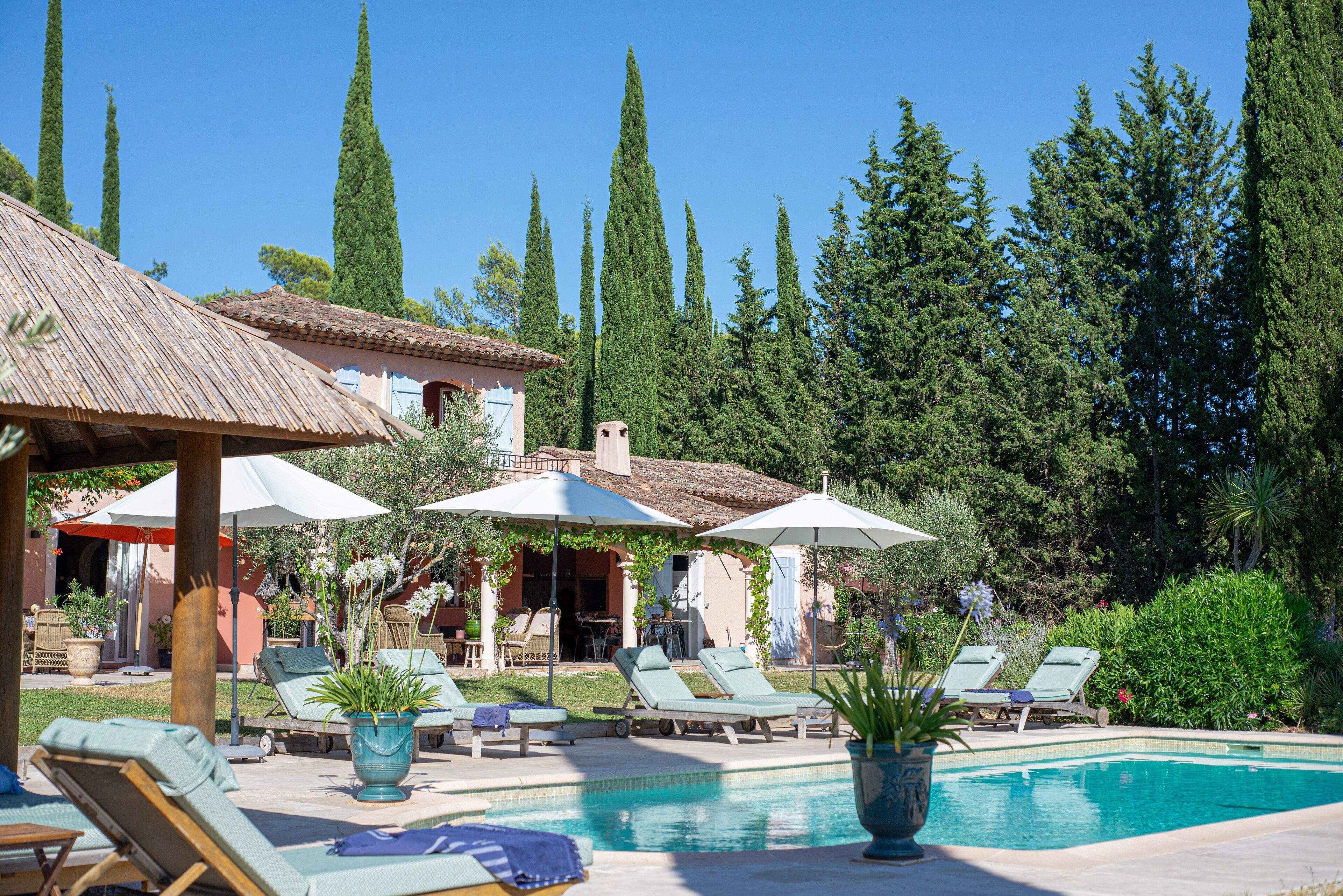 A pool area with lounge chairs and umbrellas, surrounded by tall trees and a building with a tiled roof.