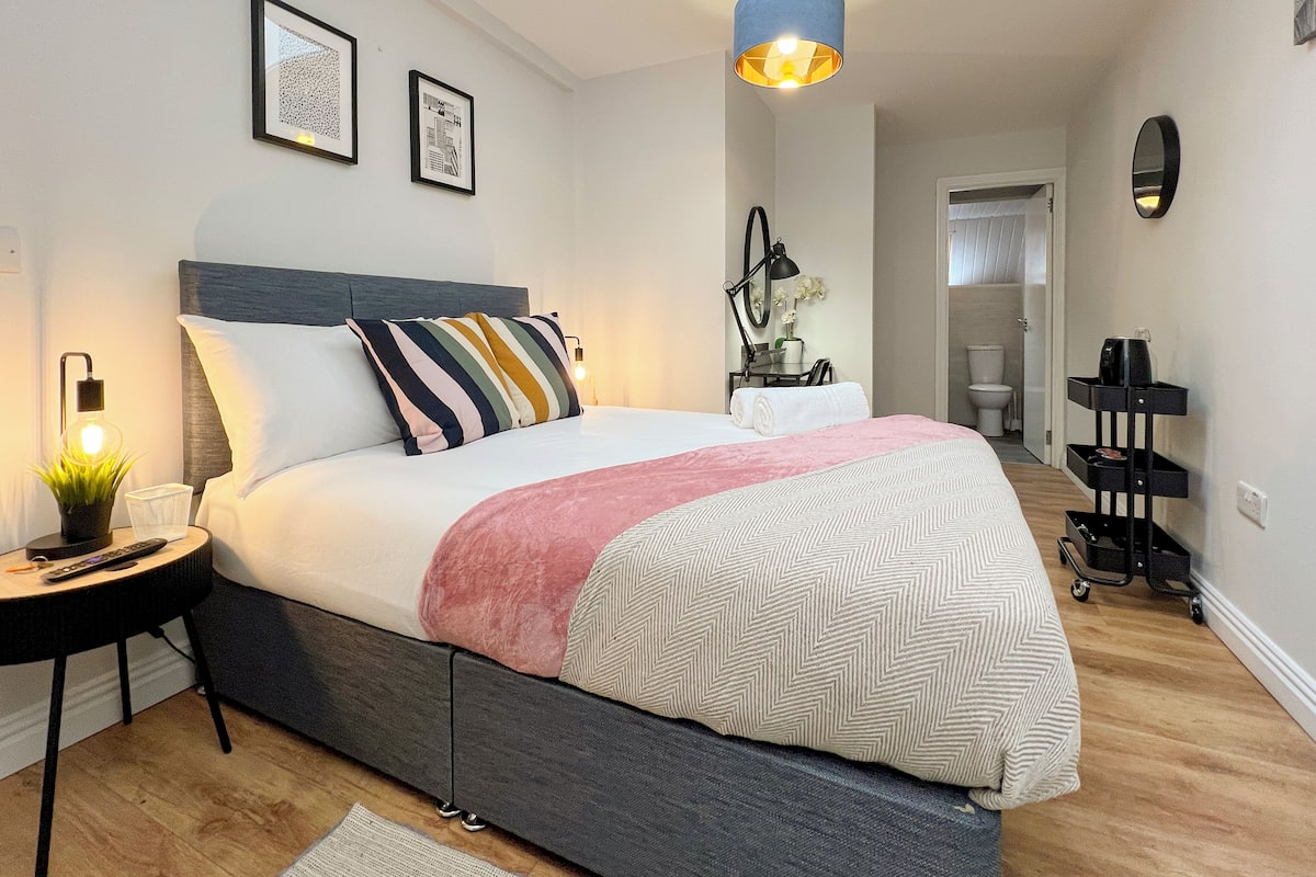 A modern bedroom with a large bed, a bedside table with a lamp, a small plant, and a wall-mounted mirror.