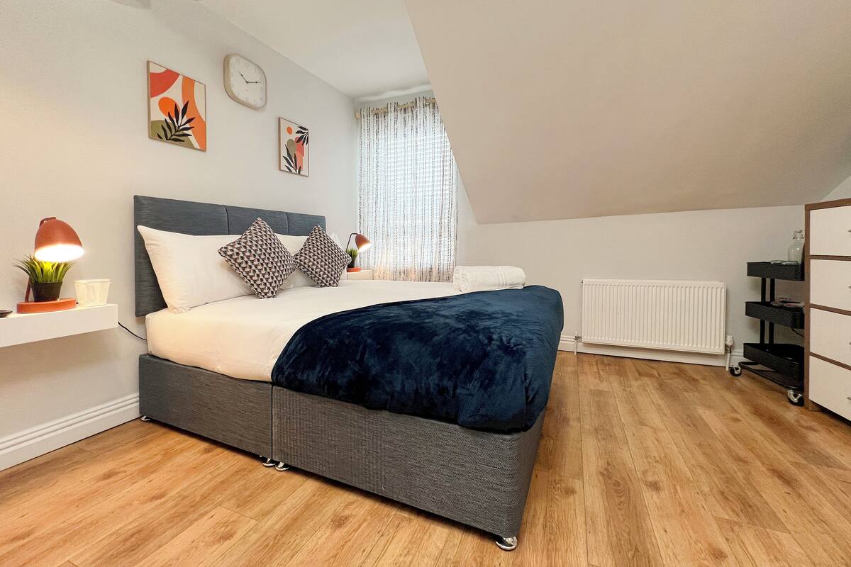 A modern bedroom with a large bed, a bedside table with a lamp, a radiator, and a window with curtains.
