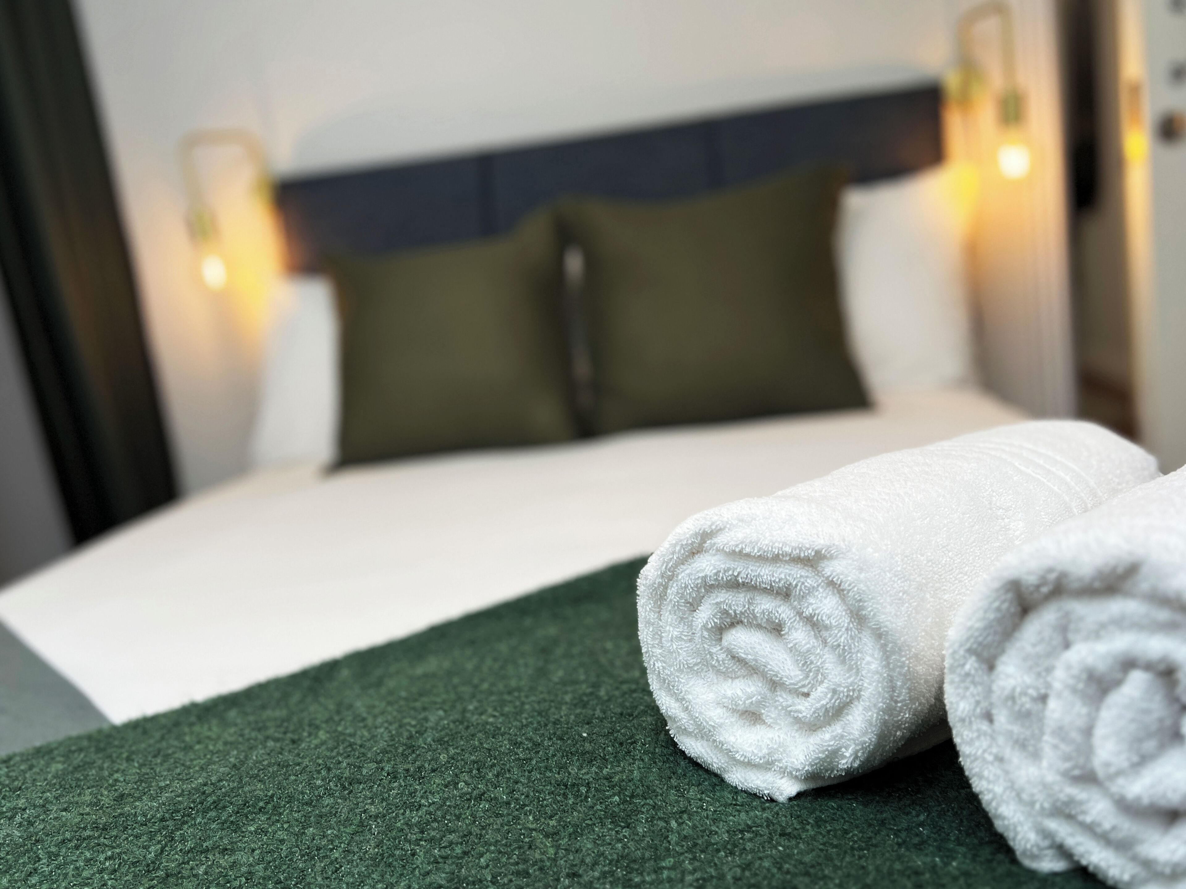 A neatly made bed with white linens, a green throw, and two rolled white towels.