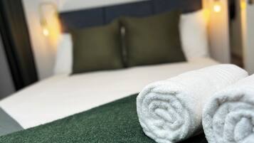 A neatly made bed with white linens, a green throw, and two rolled white towels.
