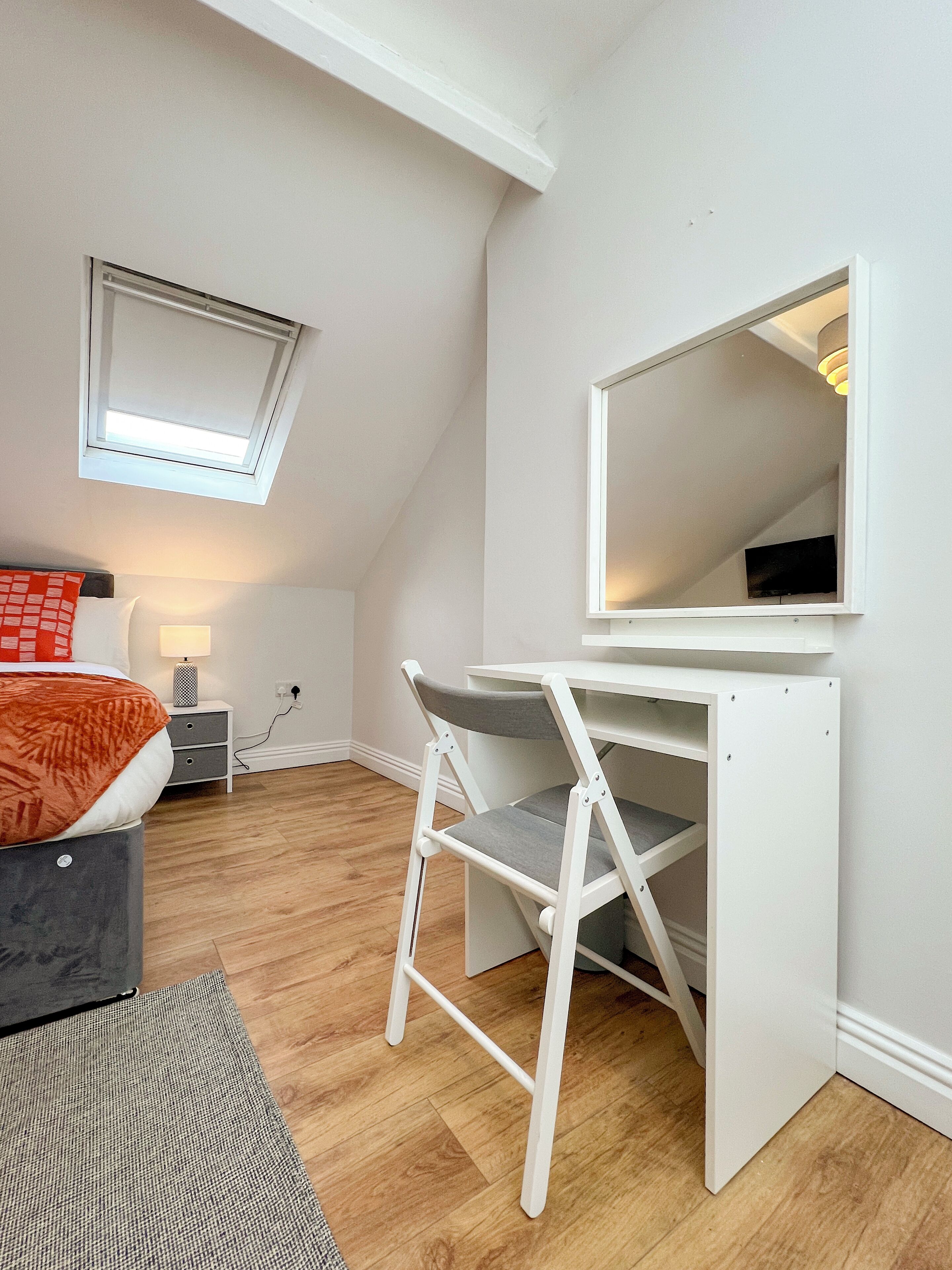 A bedroom with a bed, bedside table, lamp, and a desk with a chair. There is a skylight above the bed and a TV mounted on the wall.