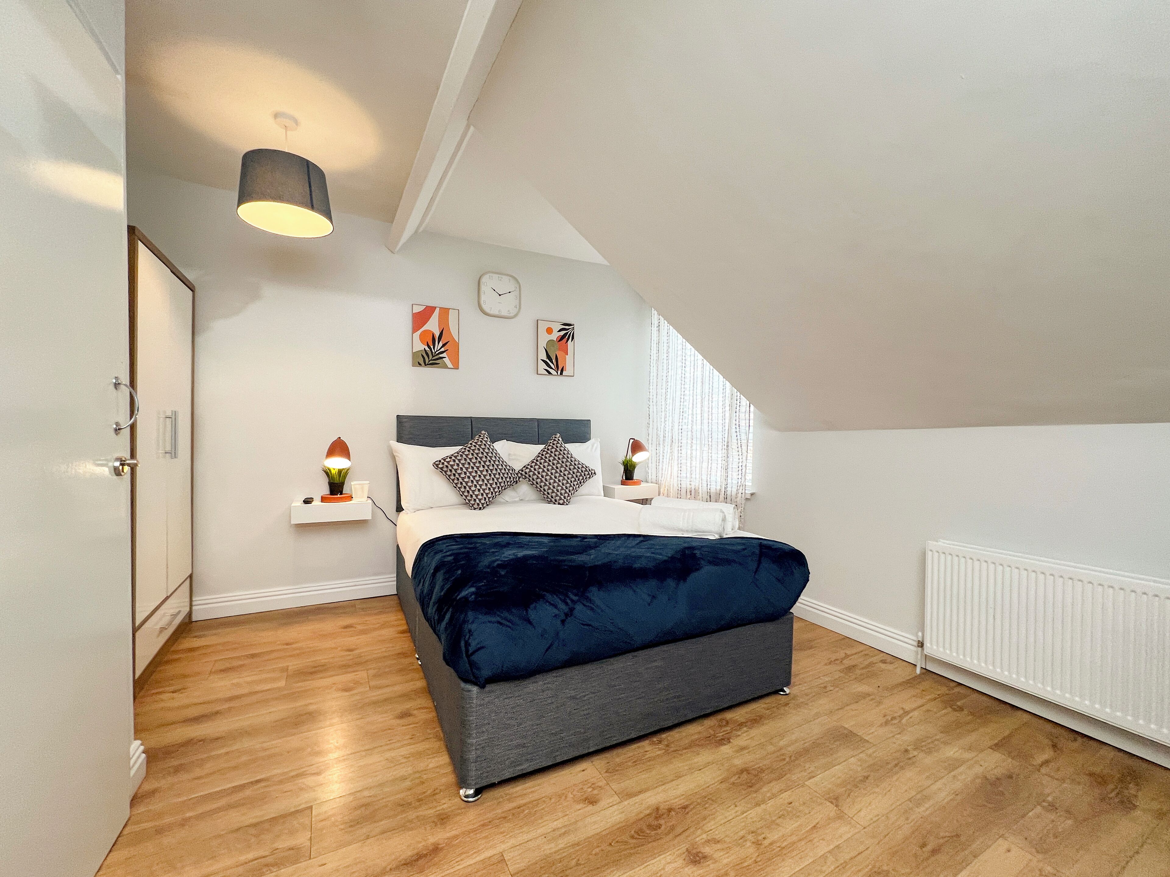 A modern bedroom with a large bed, a bedside table, a lamp, and a clock on the wall.