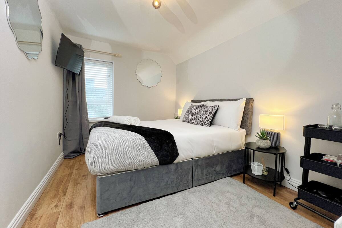 A modern bedroom with a grey bed, white bedding, a grey rug, a small bedside table with a lamp, and a wall-mounted TV.
