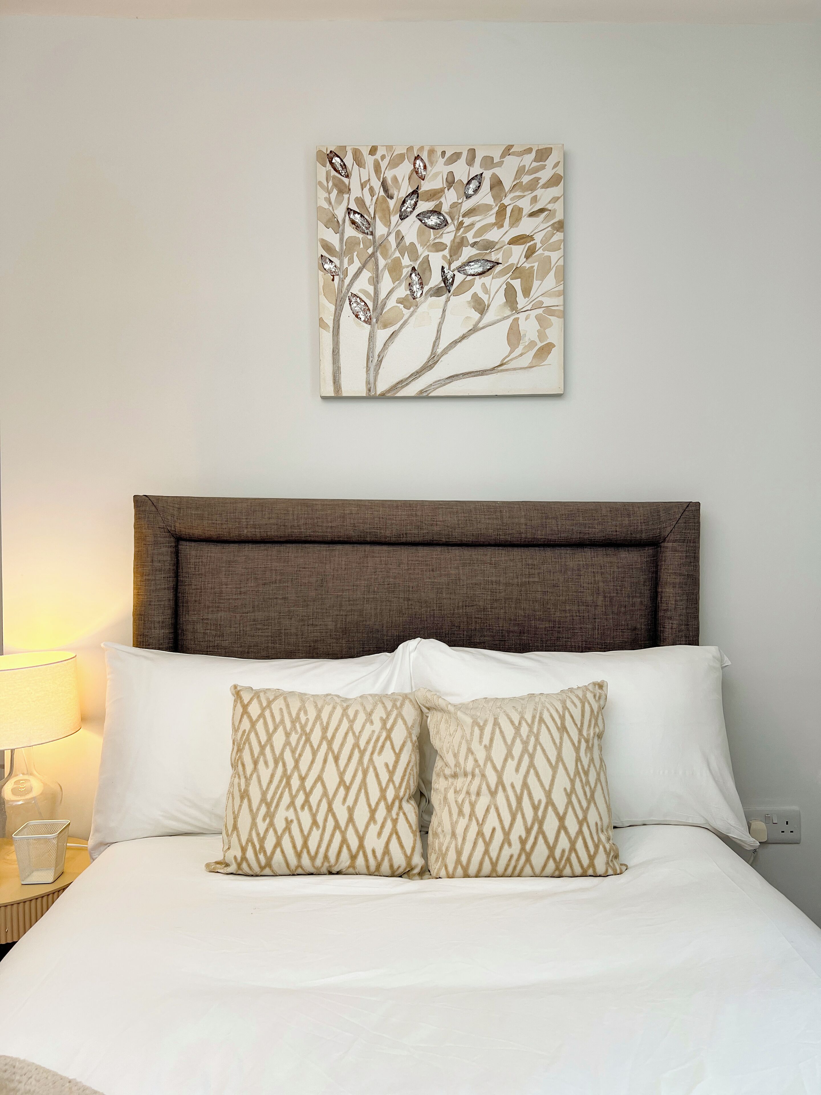 A neatly made bed with white linens and patterned pillows, a brown headboard, and a wall-mounted artwork depicting leaves.