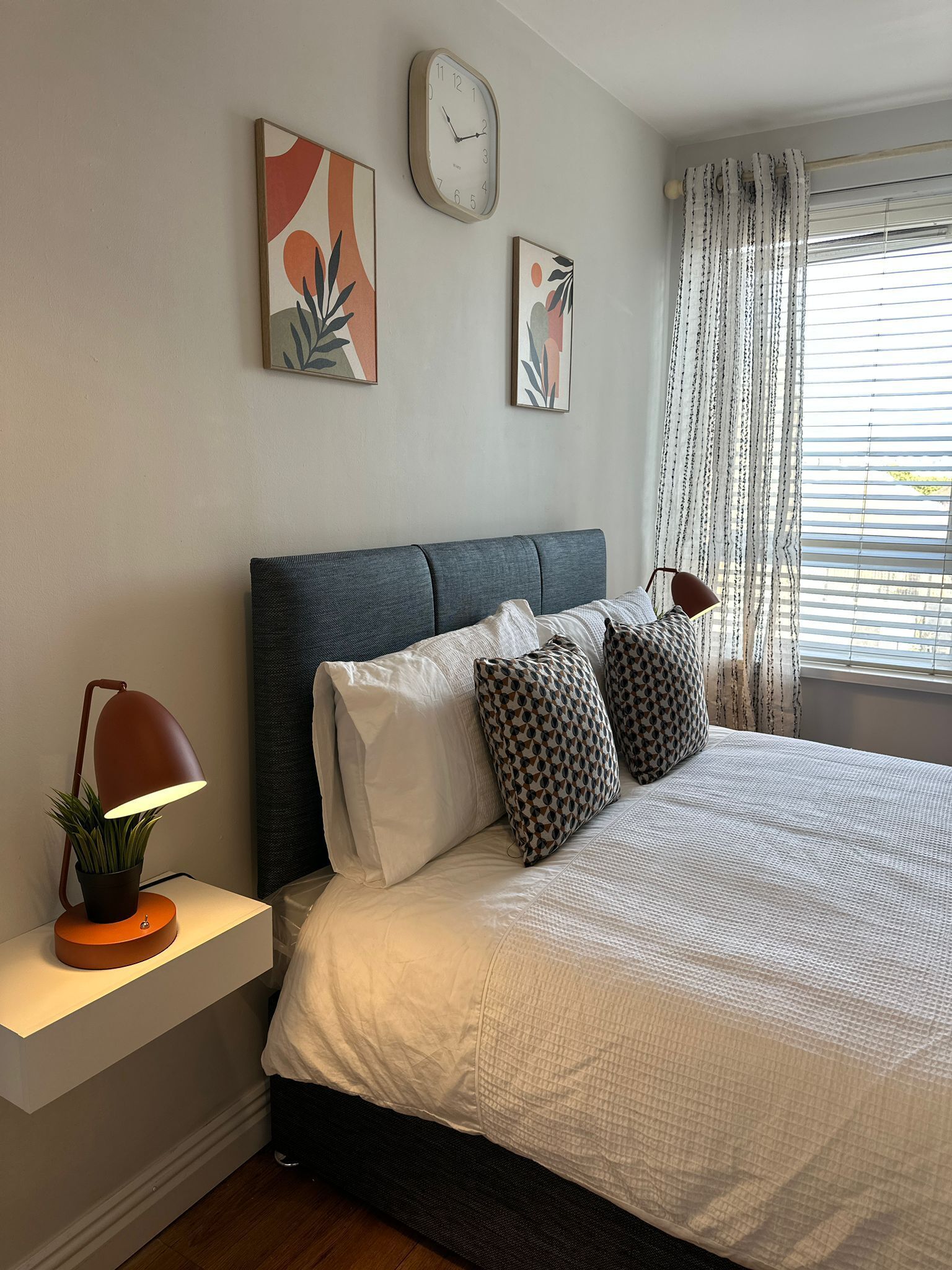A bedroom with a bed, pillows, a nightstand with a lamp, a clock, and abstract wall art.