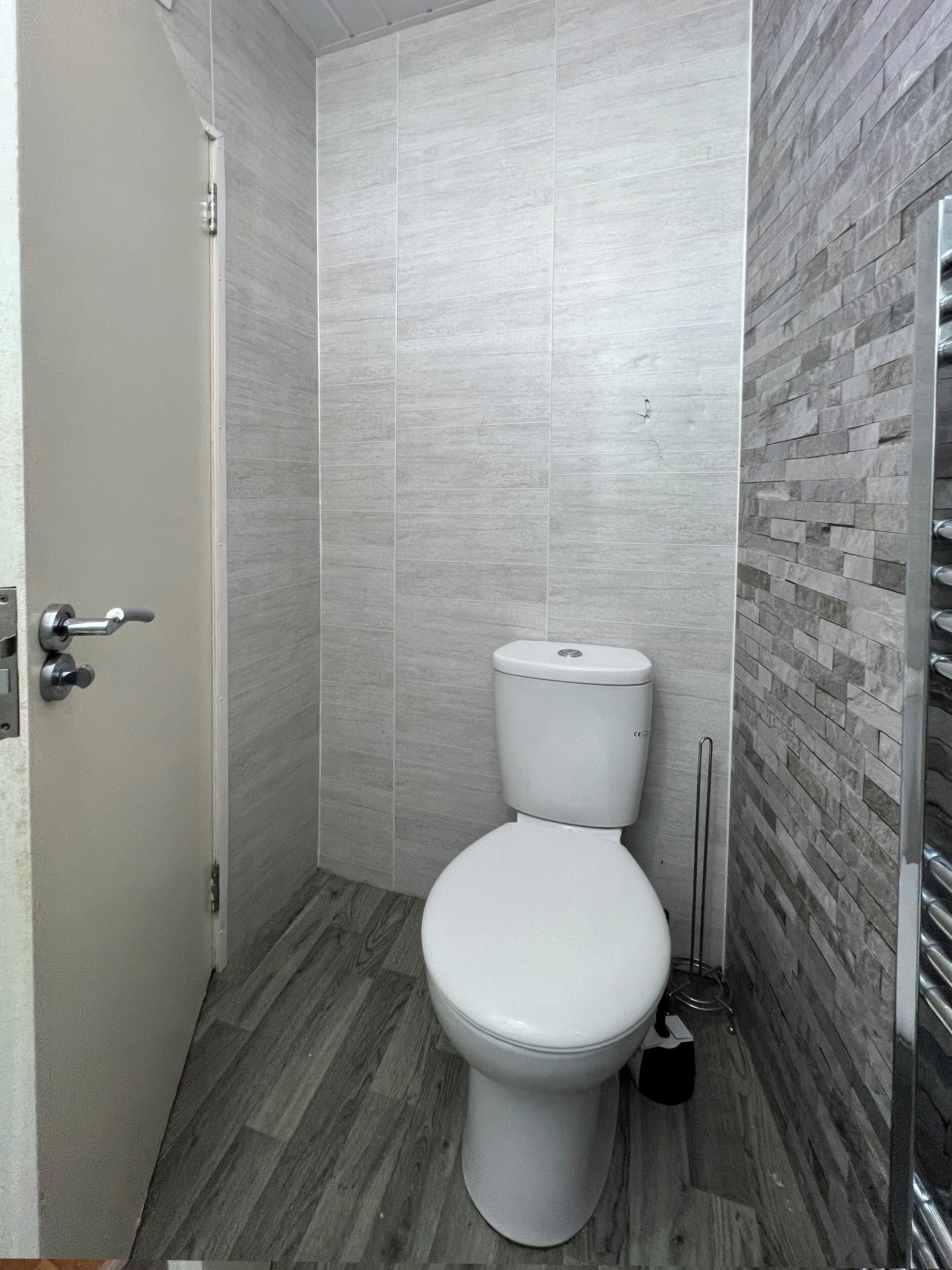 A bathroom with a white toilet, grey tiled walls, and a wooden floor.