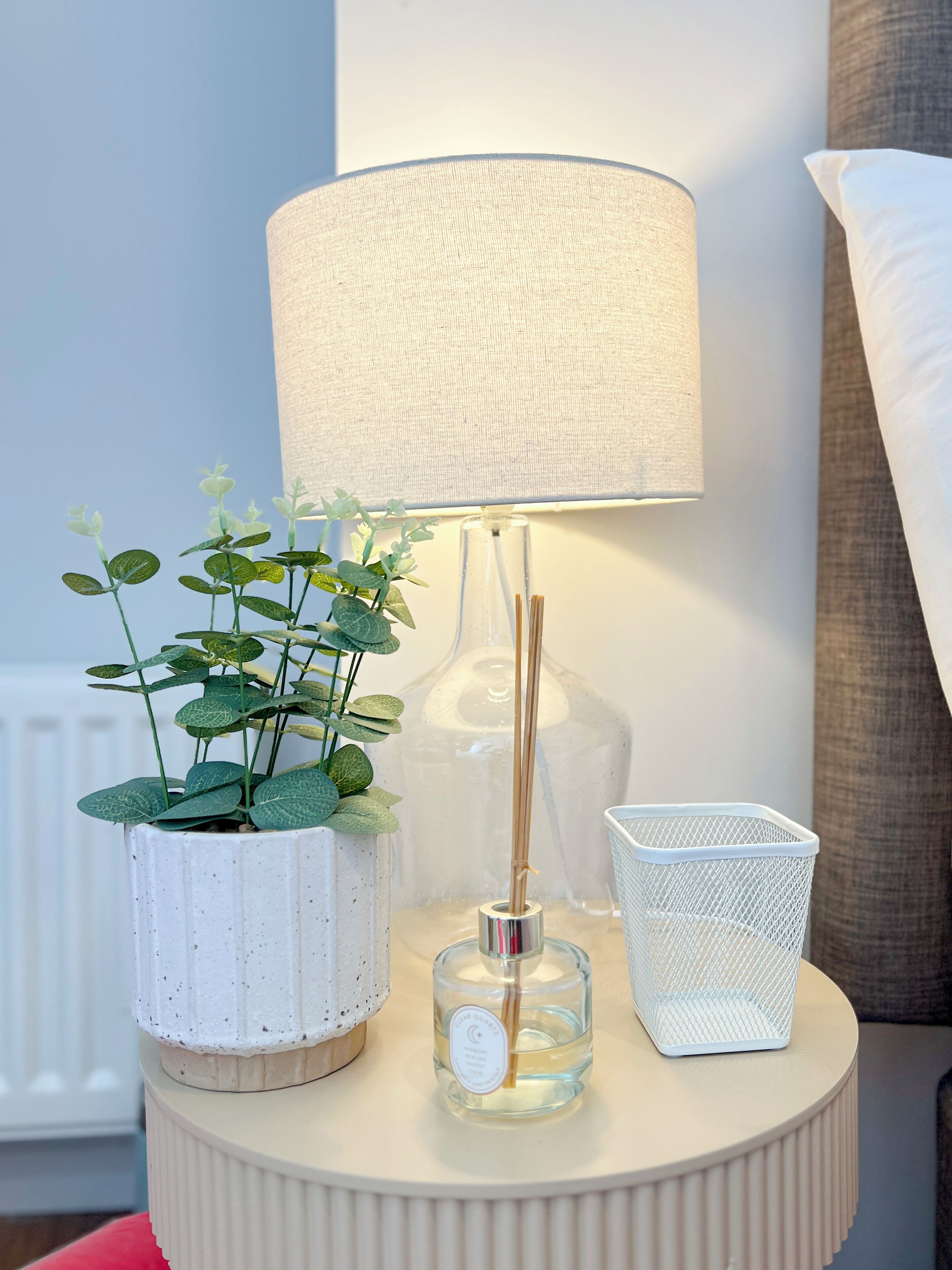 A bedside table with a lamp, a potted plant, a glass bottle with a label, and a white mesh basket.