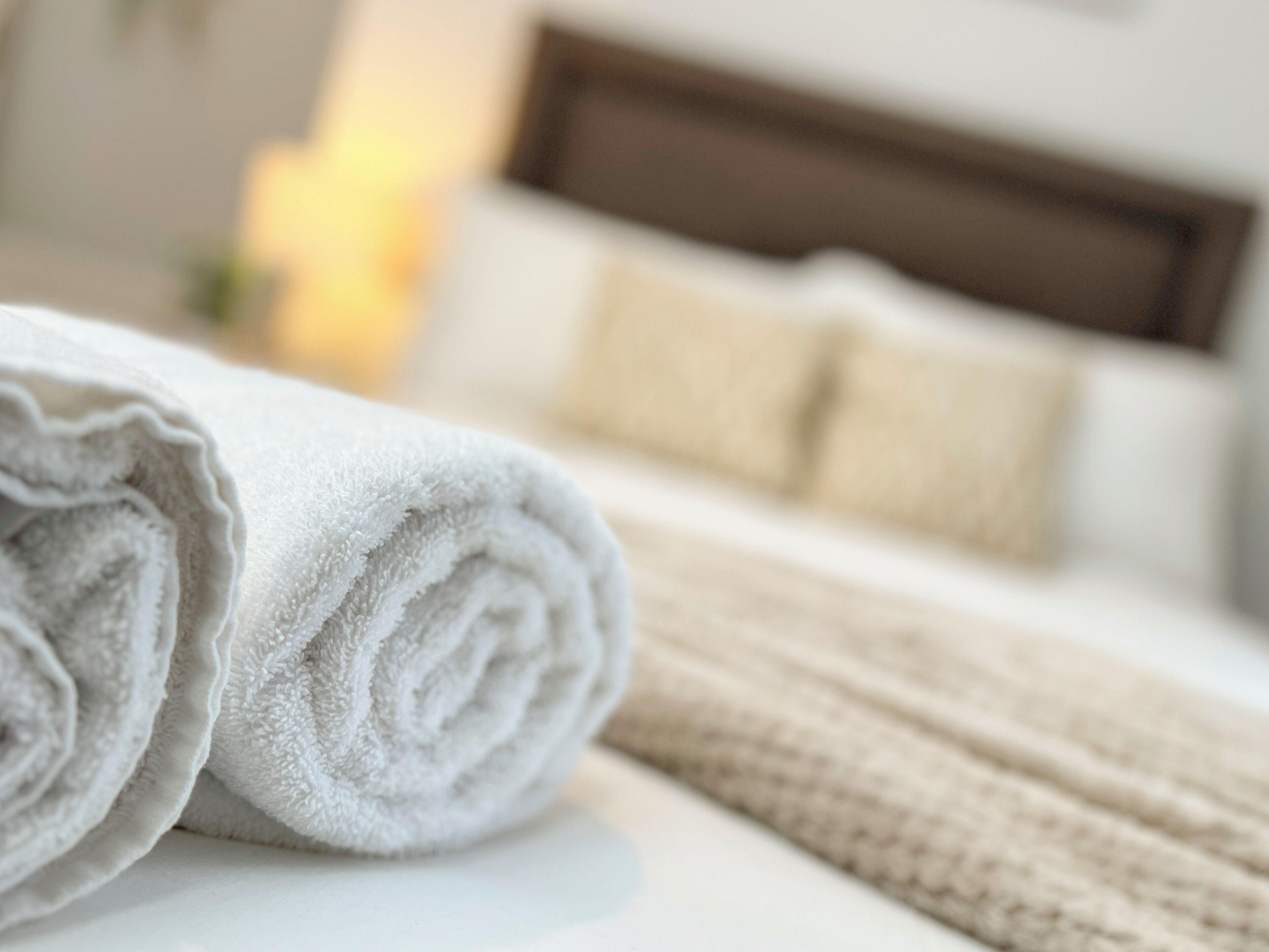 Un lit bien fait, recouvert d’une couverture blanche et beige, avec une serviette blanche enroulée et une tête de lit en bois.