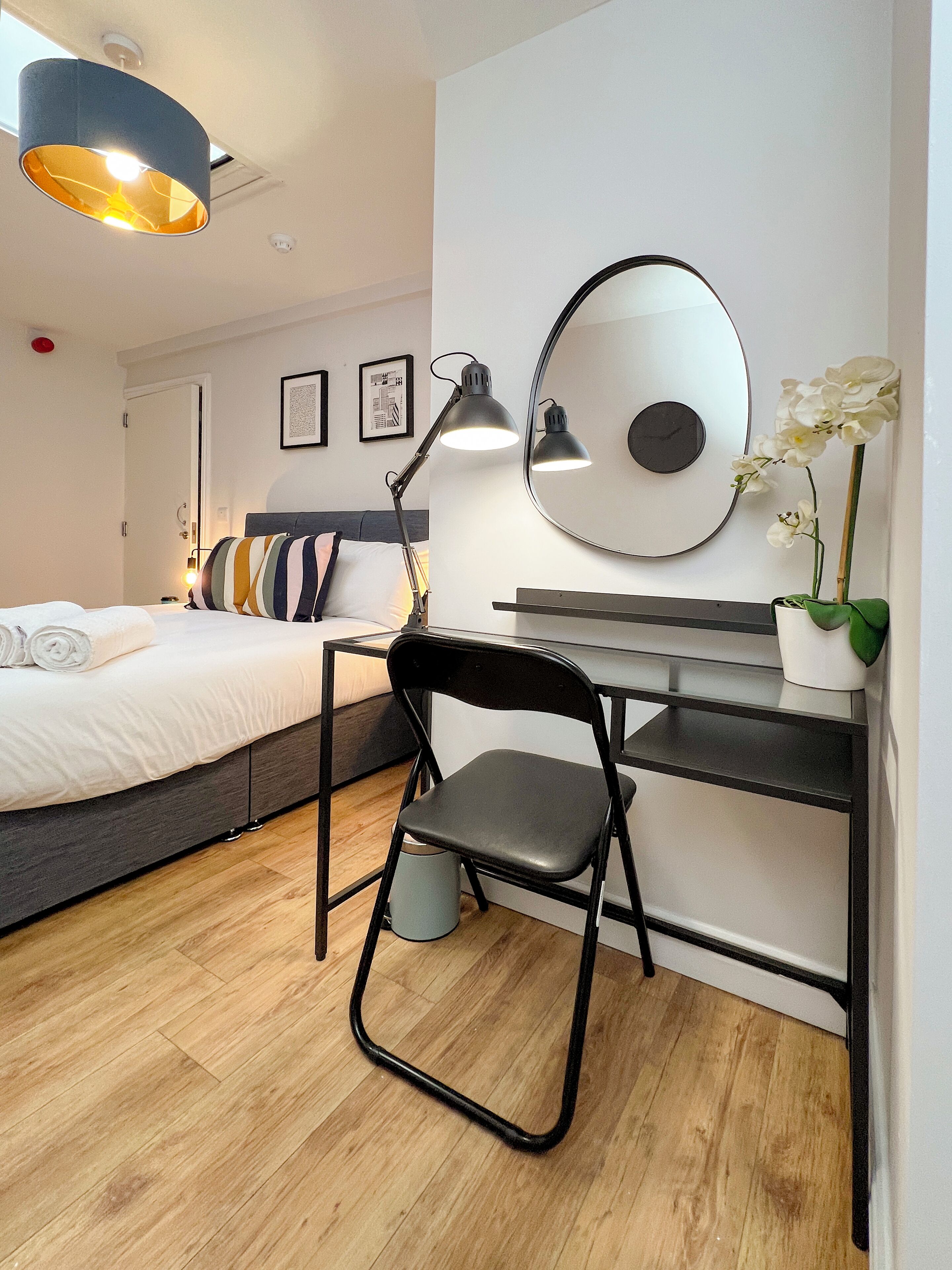 Un dormitorio moderno con cama, escritorio, silla, espejo y una orquídea en maceta.