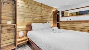 A hotel room with wooden panel walls, a bed with white linens, a bedside table with a lamp, and a mirror above the bed. - Grafton Woodshack | By My Getaways (Brighton)