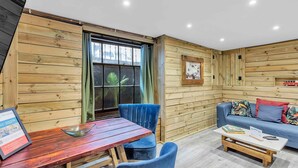 A cozy room with wooden walls, a wooden table, blue chairs, a sofa with colorful cushions, and a small shelf with a toy car. - Grafton Woodshack | By My Getaways (Brighton)
