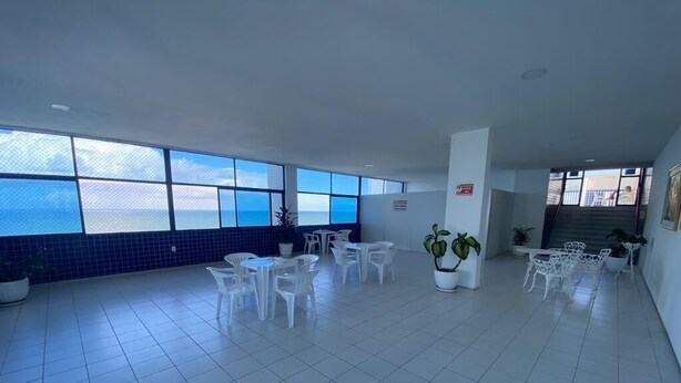 A spacious indoor area with large windows offering a view of the sea, white tiled flooring, and several white tables and chairs. - Flat Studio Everest: Your Accommodation in Boa Viagem (Recife)